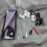 A flat-lay arrangement of a sewing and dressmaking kit on a draped light gray fabric background. The kit includes a box of Emery dressmaking scissors, three white and three black bottles of liquid nail polish with red winged logos, a red-handled clear pen, two gray pencils, a white-handled craft knife, and a small black oval sticker.
