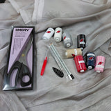 A flat-lay of sewing and embroidery supplies arranged on a light gray sheer fabric background. Visible items include multiple spools of thread in pink, navy, black, white, beige, and red, two embroidery pens, a red pen, a black oval label, a packaged pair of dressmaking scissors branded EMERY, and a small clear plastic bag with white and gray pencils.