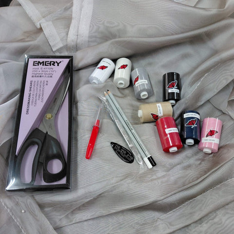 A flat-lay of sewing and embroidery supplies arranged on a light gray sheer fabric background. Visible items include multiple spools of thread in pink, navy, black, white, beige, and red, two embroidery pens, a red pen, a black oval label, a packaged pair of dressmaking scissors branded EMERY, and a small clear plastic bag with white and gray pencils.